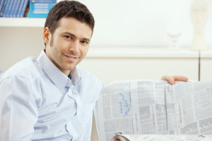 Casual young businessman reading business news, sitting on couch at home, looking at camera, smiling.